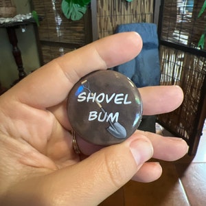 May include: A brown button with a white shovel and the text "SHOVEL BUM" on it.