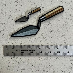 Puede incluir: Dos parches de paleta de hierro bordados, uno m&aacute;s grande que el otro, con un mango de madera marr&oacute;n y una hoja de metal gris. Los parches est&aacute;n dispuestos sobre una encimera blanca con una regla para la escala.