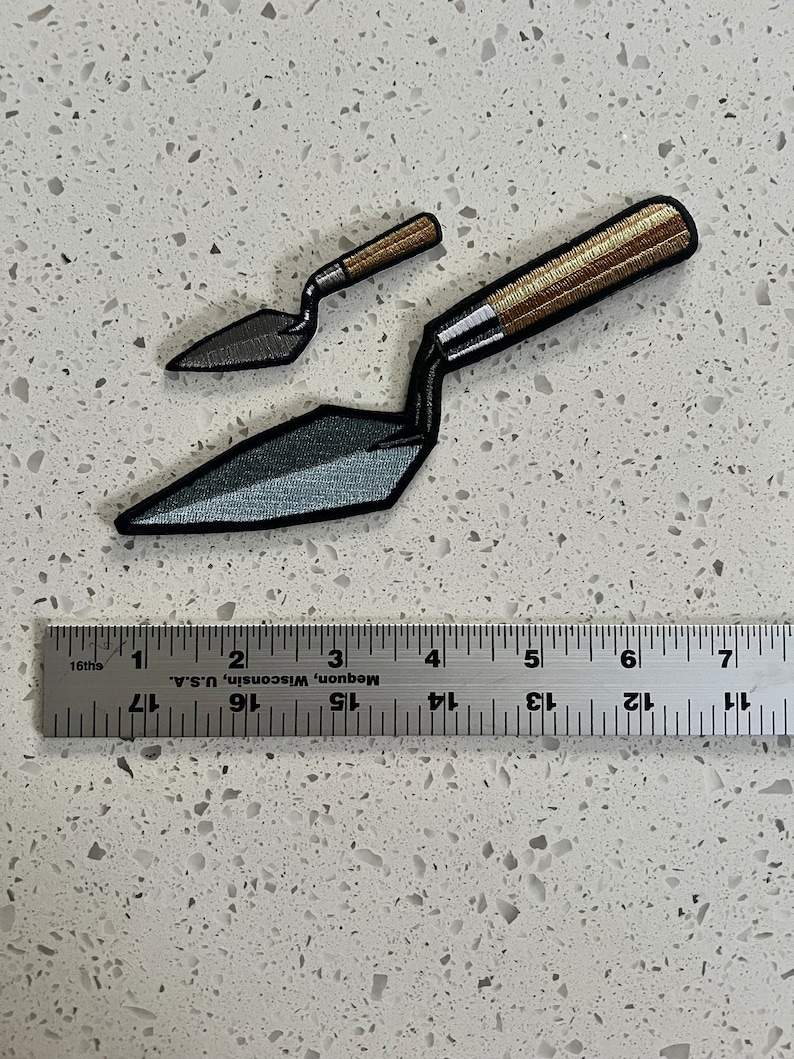 Puede incluir: Dos parches de paleta de hierro bordados, uno m&aacute;s grande que el otro, con un mango de madera marr&oacute;n y una hoja de metal gris. Los parches est&aacute;n dispuestos sobre una encimera blanca con una regla para la escala.