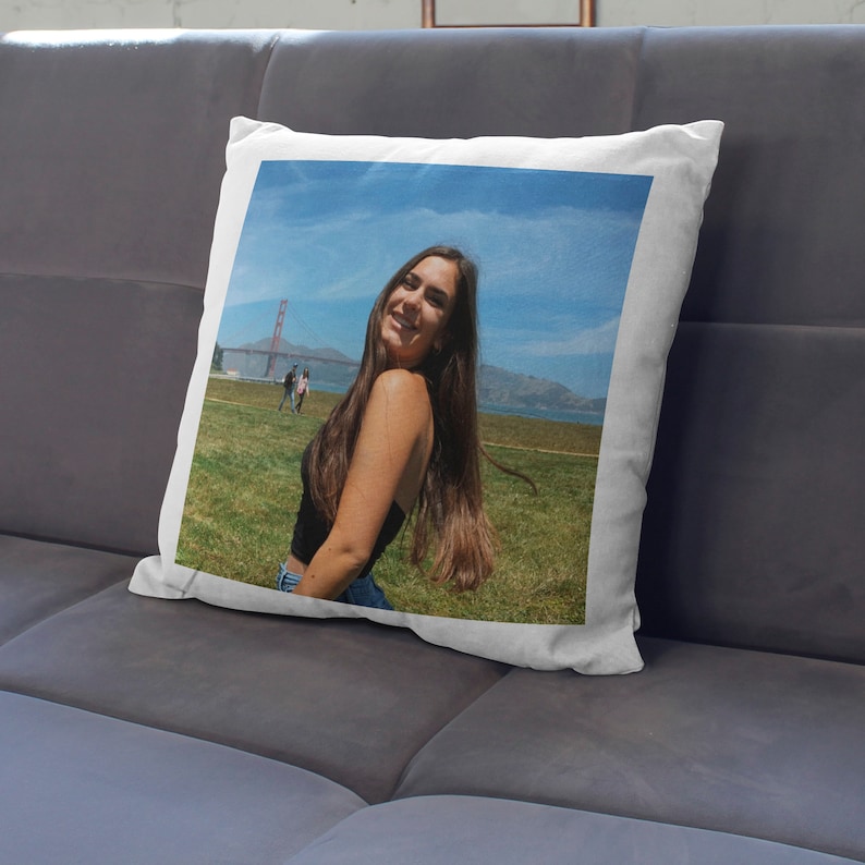 May include: Square white cushion featuring a photograph of a person smiling in front of the Golden Gate Bridge. The image shows a blue sky, green grass, and the iconic red bridge. The cushion is on a grey sofa.