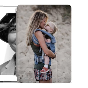 May include: A woman wearing a patterned skirt and a blue and grey baby carrier is holding a baby. The baby is wearing sunglasses and looking at the woman. The background is a sandy beach.