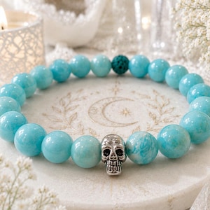 Op de afbeelding: Een kralenarmband met gladde, ronde, lichtblauwe edelsteenkralen, één enkele getextureerde donkergroene kraal en een centrale schedelbedel van zilverkleurig metaal. De kralen zijn geregen aan een elastisch koord in een cirkelvormig ontwerp.