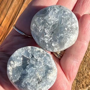 May include: Two light blue, rounded celestite crystals. The crystals have a rough, textured surface and a pale blue color. They are held in a person's hand, with a wooden surface in the background. The crystals are a natural, mineral product.