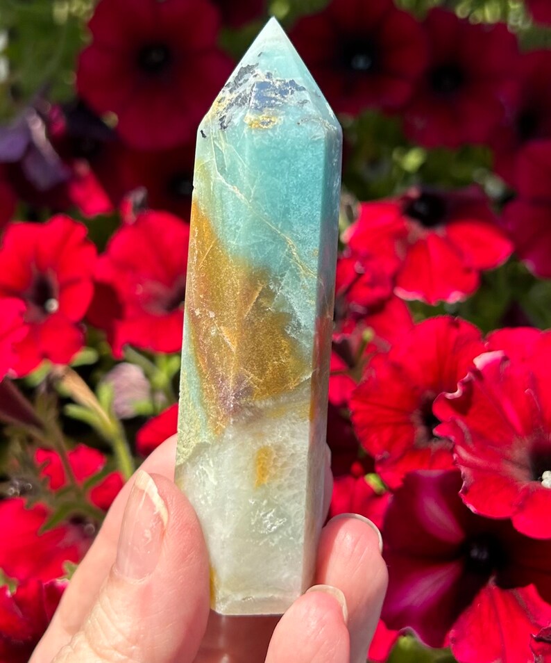 May include: A polished crystal obelisk with a pointed top. The crystal features a blend of colors, including light blue, gold, and white. The obelisk is held in front of a backdrop of vibrant red flowers.