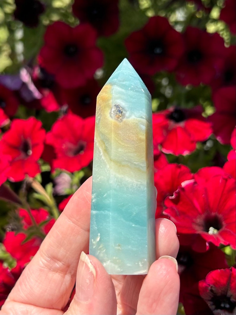 May include: A polished crystal tower with a light blue and tan color scheme. The crystal has a pointed top and is held in a person's hand. The background features red flowers.