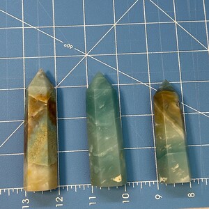 May include: Three polished crystal obelisks in varying sizes. The crystals display a mix of colors, including green, brown, and cream. They are set against a blue grid background, with measurements in inches visible.