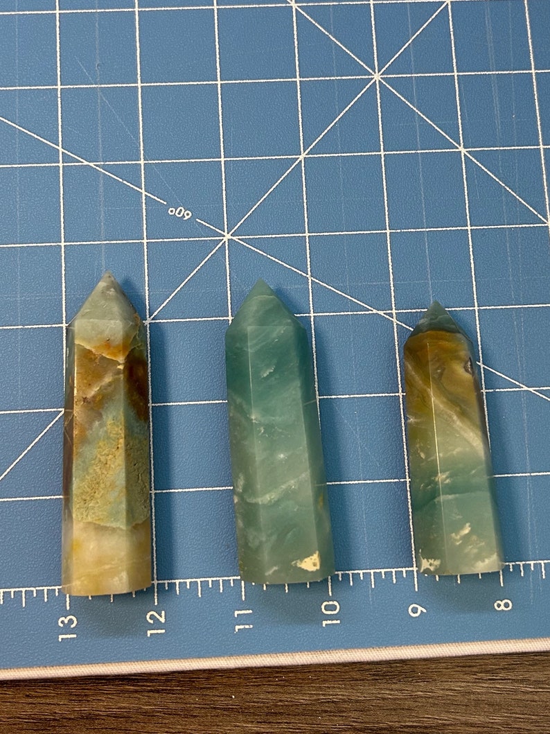 May include: Three polished crystal obelisks in varying sizes. The crystals display a mix of colors, including green, brown, and cream. They are set against a blue grid background, with measurements in inches visible.