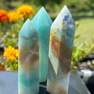 May include: Three polished crystal towers in shades of blue, brown, and white. The crystals have a rectangular base and a pointed top. The background features blurred yellow and orange flowers and green foliage.