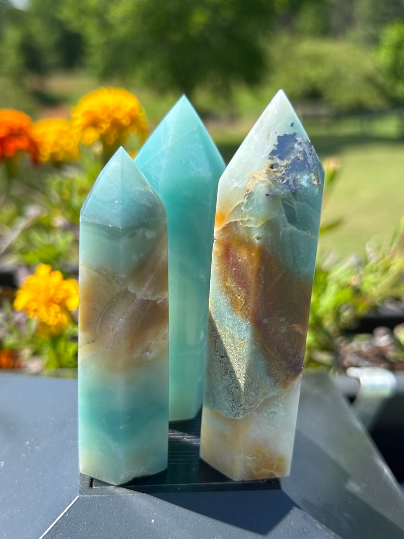 May include: Three polished crystal towers in shades of blue, brown, and white. The crystals have a rectangular base and a pointed top. The background features blurred yellow and orange flowers and green foliage.