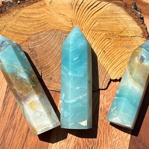 May include: Three polished crystal towers in shades of blue, teal, and tan are displayed on a wooden surface. Each tower has a rectangular base and tapers to a point. The crystals have a translucent quality, with visible internal patterns.