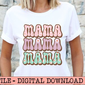 May include: White t-shirt with the word "MAMA" in three rows, each in a different color and design. The top row is pink with white flowers, the middle row is purple with white flowers, and the bottom row is teal with white flowers.
