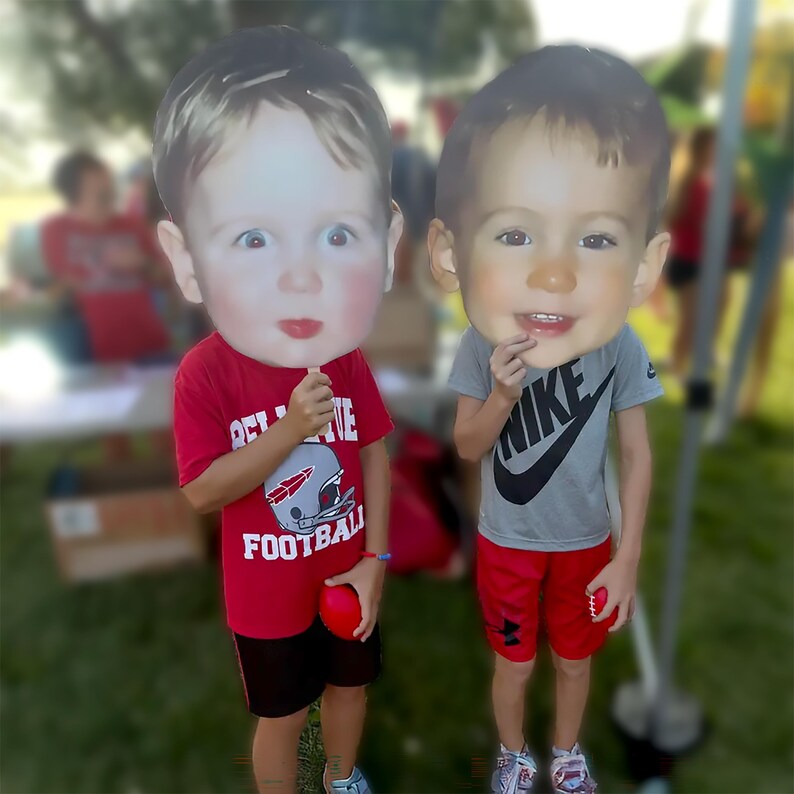Peut inclure: Deux grandes d&eacute;coupes en carton de visages d'enfants tenues sur des b&acirc;tons. Un enfant porte un t-shirt rouge avec un graphique de football et du texte, tenant une balle rouge. L'autre porte un t-shirt Nike gris et un short rouge, tenant un objet rouge.