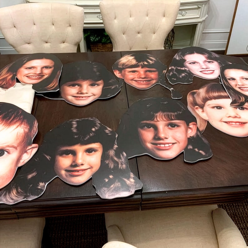Peut inclure: Une collection de huit d&eacute;coupes de visage en carton mousse, imprim&eacute;es sur mesure, repr&eacute;sentant divers portraits d'enfants et d'adultes avec des coiffures et des expressions vari&eacute;es, dispos&eacute;es sur une surface en bois sombre.