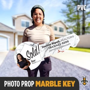 May include: A white marble key-shaped photo prop with the words "Sold!" and "kw KELLERWILLIAMS" on it. The key also has a business card with the name "Jasmin Ripoll, REALTOR®", phone number, and email address. The prop is being held by a person wearing a white shirt and black pants.