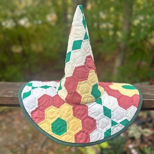 May include: A handmade, quilted hat with a wide brim and a tall, pointed crown. The hat features a patchwork design of red, yellow, green, and white geometric shapes. The brim is edged with green fabric. The hat is likely for costume or decorative purposes.