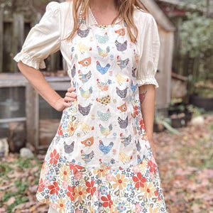 May include: A white apron with a colourful chicken pattern and a floral border. The apron features a variety of chicken illustrations in different colours and patterns. The bottom edge is adorned with a vibrant floral print in shades of red, orange, blue, and yellow.