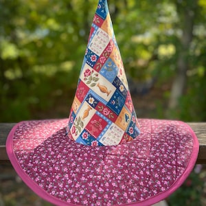 May include: A quilted witch hat with a wide brim. The cone-shaped crown is patchwork with floral and geometric patterns in red, blue, yellow, and white. The brim is a deep pink with a floral print and a pink trim.