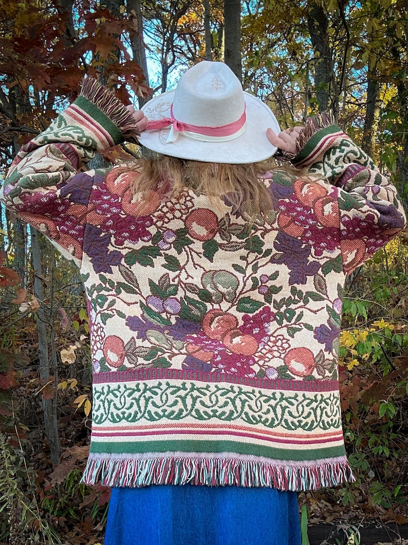 May include: A tapestry jacket with a floral pattern in shades of burgundy, green, and peach. The jacket has a fringed hem and sleeves. A white hat with a pink ribbon is also visible.