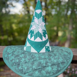 May include: A handmade witch hat with a wide brim. The hat is made of quilted fabric in shades of green and white. The conical crown features a starburst pattern. The brim has a floral design and is edged with a darker green binding.