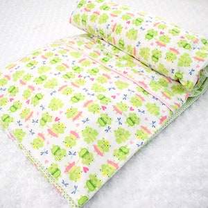 May include: A white and green baby blanket with a pattern of frogs wearing pink tutus and crowns. The blanket is folded in half and has a pink trim.