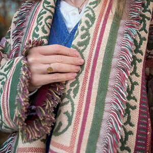 May include: A woven shawl with a colorful pattern. The shawl features a repeating green leaf design, alternating with stripes of beige, red, and green. The edges are finished with a fringe in shades of green, purple, and beige.