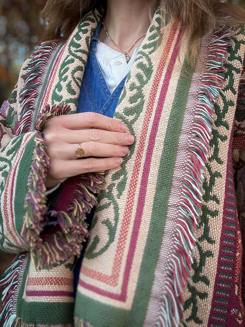May include: A woven shawl with a colorful pattern. The shawl features a repeating green leaf design, alternating with stripes of beige, red, and green. The edges are finished with a fringe in shades of green, purple, and beige.