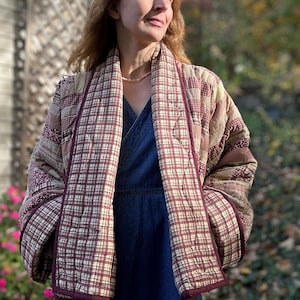May include: A quilted jacket featuring a patchwork design with various plaid and floral patterns in shades of burgundy, tan, and cream. The jacket has a shawl collar, long sleeves, and a dark burgundy trim along the open front edges.