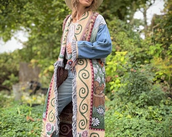 May include: A long, beige tapestry coat with a denim jacket overlay. The coat features a floral and scroll design in shades of brown, green, and burgundy, with a fringed hem. The model is wearing a wide-brimmed hat, light blue jeans, and beige boots.