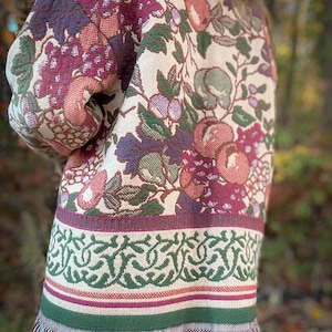 May include: A tapestry jacket with a cream background and a floral pattern featuring grapes, apples, and leaves in shades of purple, pink, and green. The jacket has a green and cream border with fringe at the bottom.