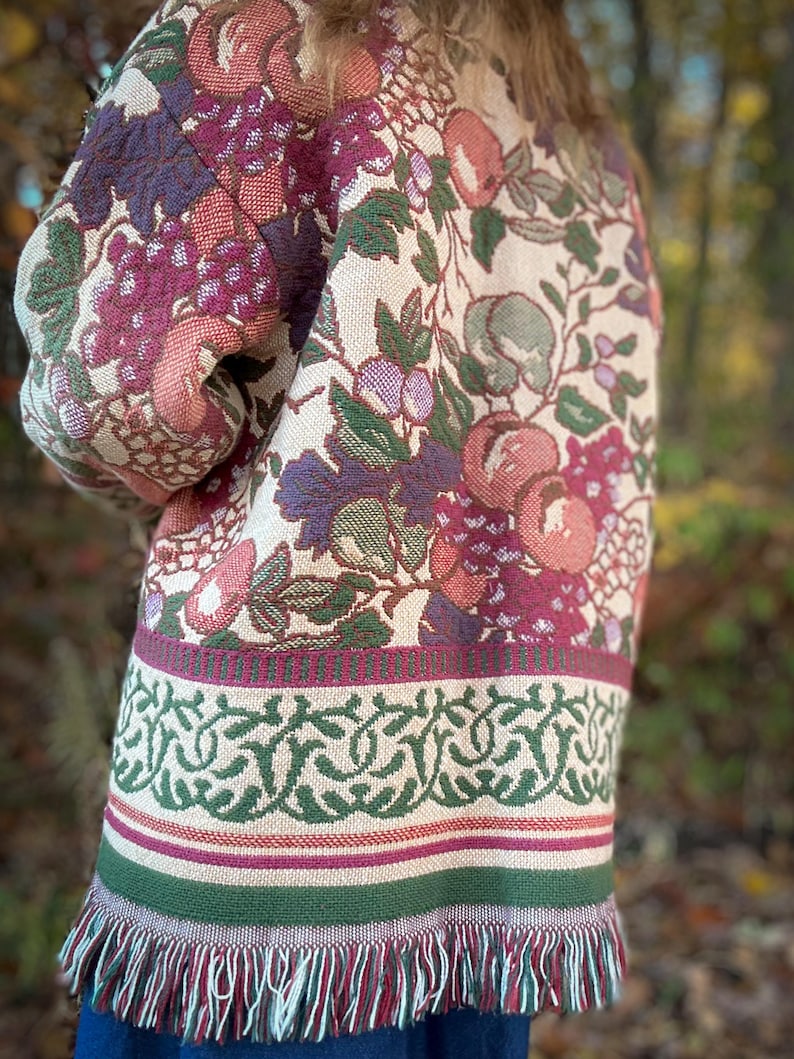 May include: A tapestry jacket with a cream background and a floral pattern featuring grapes, apples, and leaves in shades of purple, pink, and green. The jacket has a green and cream border with fringe at the bottom.
