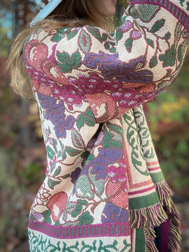 May include: A woven tapestry shawl with a floral pattern in shades of cream, green, purple, and red. The shawl features a fringed edge and a repeating botanical design. The fabric appears to be thick and textured, suitable for cooler weather.