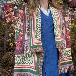 May include: A tapestry-style cardigan with a floral and foliage pattern in shades of cream, burgundy, green, and brown. The open-front cardigan has a fringed hem and sleeves. The garment is worn over a denim dress.