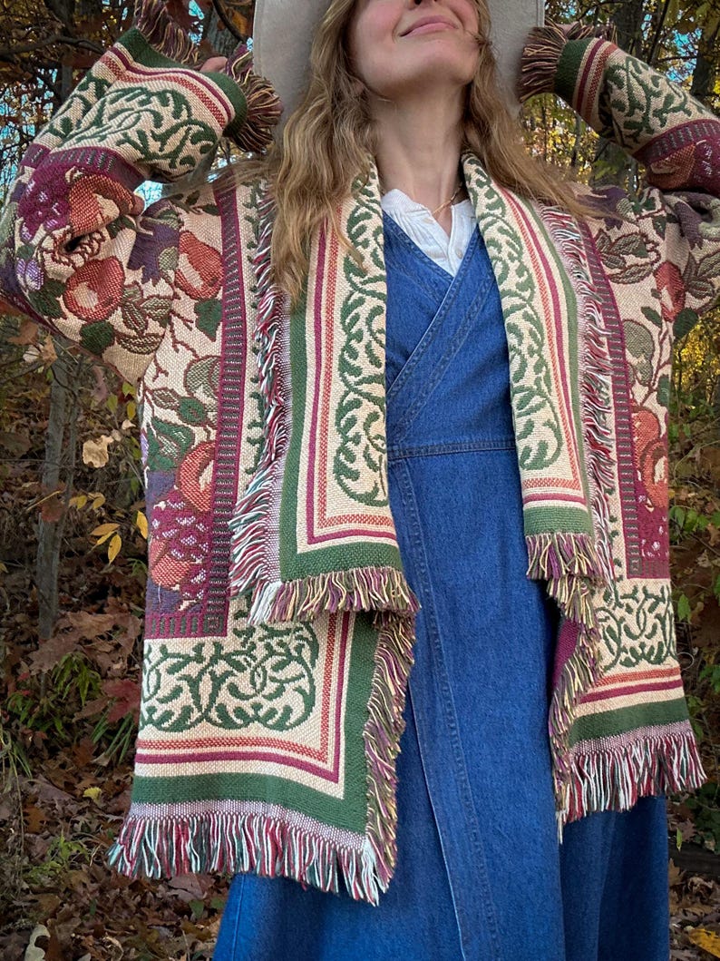 May include: A tapestry-style cardigan with a floral and foliage pattern in shades of cream, burgundy, green, and brown. The open-front cardigan has a fringed hem and sleeves. The garment is worn over a denim dress.