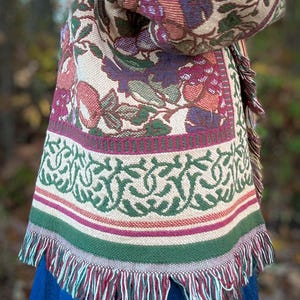 May include: A woven shawl with a floral pattern in shades of burgundy, green, and beige. The shawl has a fringed edge and a decorative border with a green and white design. The shawl is draped over a blue garment.