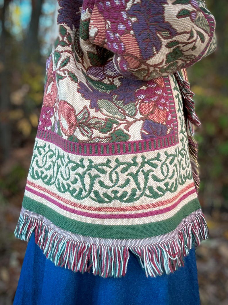May include: A woven shawl with a floral pattern in shades of burgundy, green, and beige. The shawl has a fringed edge and a decorative border with a green and white design. The shawl is draped over a blue garment.