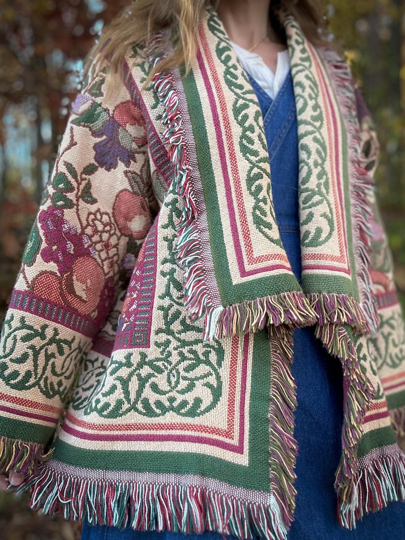 May include: A tapestry jacket with a floral and geometric pattern in shades of beige, green, burgundy, and purple. The jacket has a fringed hem and edges. The design includes botanical and ornamental elements.