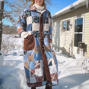 May include: A long patchwork coat featuring a mix of blue, white, and patterned fabrics. The coat has a brown suede-like panel down the front with button closures and large pockets. The coat is worn over a white turtleneck in a snowy outdoor setting.