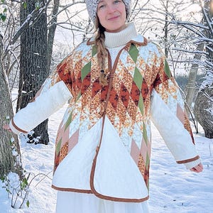 May include: A white quilted jacket with a geometric pattern in shades of brown, green, and pink. The jacket has a button closure and is worn over a white turtleneck sweater. The person is wearing a gray knitted hat and is standing in a snowy forest.