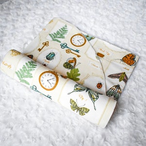 May include: A beige fabric with a repeating pattern of butterflies, keys, clocks, and insects. The fabric is rolled up and the pattern is visible on both sides of the roll.