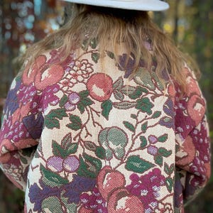 May include: A person wearing a cream-colored hat with a pink ribbon and a floral tapestry jacket. The jacket features a cream background with a pattern of red, purple, and green flowers and foliage.