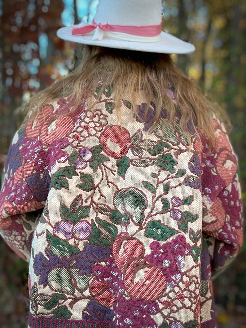 May include: A person wearing a cream-colored hat with a pink ribbon and a floral tapestry jacket. The jacket features a cream background with a pattern of red, purple, and green flowers and foliage.