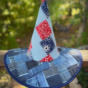 May include: A patchwork witch hat with a denim brim and a cone-shaped crown. The crown features red paisley, blue gingham, and navy blue patterned fabric. The brim is made of various denim patches, with a dark blue trim.