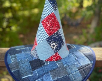 May include: A patchwork witch hat with a denim brim and a cone-shaped crown. The crown features red paisley, blue gingham, and navy blue patterned fabric. The brim is made of various denim patches, with a dark blue trim.