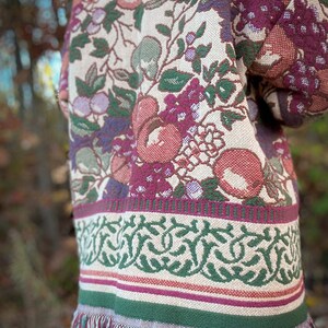 May include: A tapestry jacket with a floral pattern in shades of red, green, and purple. The jacket has a fringed hem and a decorative border. The design features apples, grapes, and leaves.