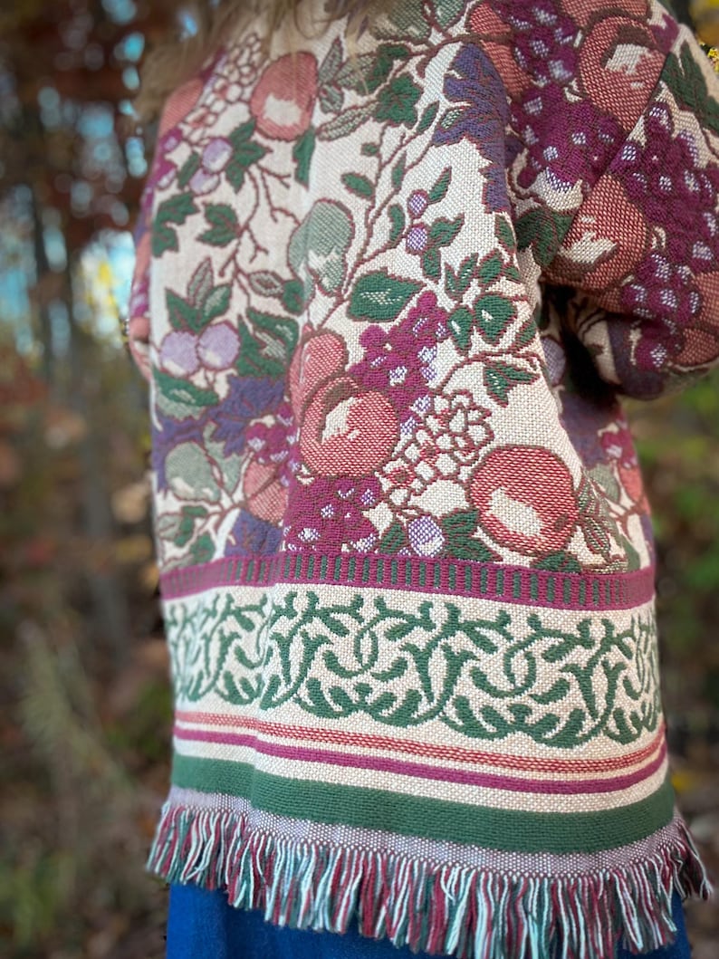 May include: A tapestry jacket with a floral pattern in shades of red, green, and purple. The jacket has a fringed hem and a decorative border. The design features apples, grapes, and leaves.