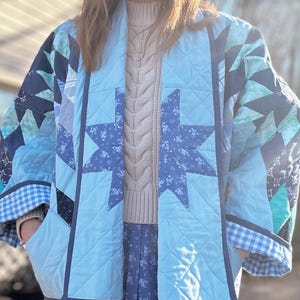 May include: A light blue quilted jacket with a star pattern. The jacket has a shawl collar and a gingham check pattern on the cuffs. The jacket is worn over a cream cable knit sweater and a floral print skirt.