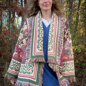 May include: A tapestry jacket with a floral and geometric pattern in shades of beige, green, and burgundy. The jacket has a fringed hem and borders. The person is wearing a denim dress and a wide-brimmed hat.