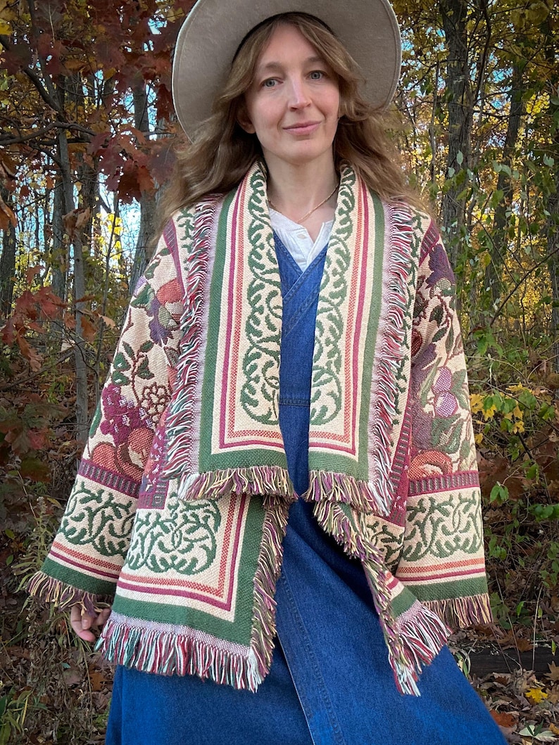 May include: A tapestry jacket with a floral and geometric pattern in shades of beige, green, and burgundy. The jacket has a fringed hem and borders. The person is wearing a denim dress and a wide-brimmed hat.