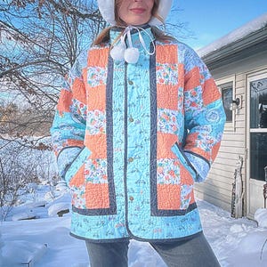 May include: A quilted jacket with a patchwork design in shades of coral, light blue, and floral patterns. The jacket has a button-up front, pockets, and a dark gray trim. The person is wearing a white hat with pom-poms.