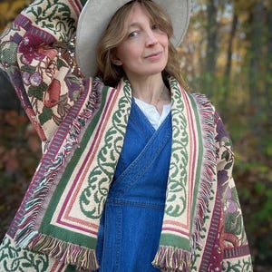 May include: A beige hat and a patterned shawl with green, burgundy, and beige floral and geometric designs. The shawl has a fringed edge. The person is wearing a blue denim dress.